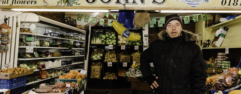 A middle aged white man wearing a black coat and woolly hat stands in a grocer’s shop amongst fruit and veg with a sign saying ‘John’s’ above him.