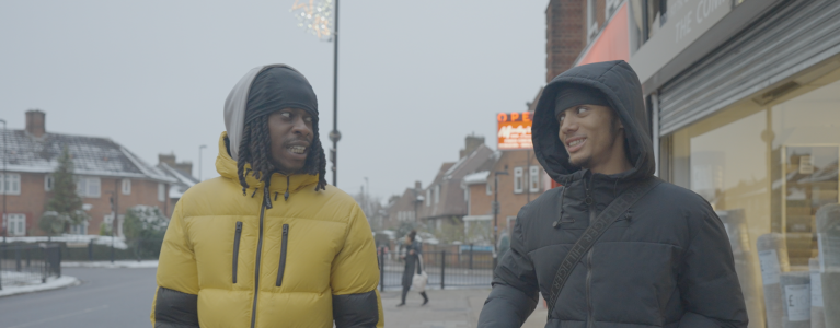 Two young men walk side by side in the street, wearing puffer ja.ckets and hoods, they look at each other and smile