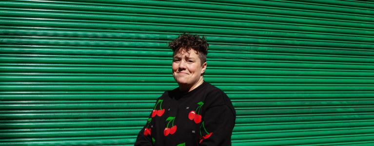 This is a picture of Jess Thom – a white woman with curly brown hair sitting in her wheelchair in front of some metallic green shop shutters on a sunny afternoon. Jess is smiling at the camera and has her hands together in her lap. She is wearing a black tracksuit with a colourful red cherry repeat print and black trainers.