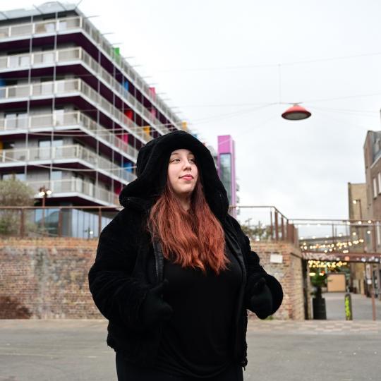 A young white woman wearing a large black hooded jacket, she looks in the distance, a backdrop of buildings and urban bridges and structures