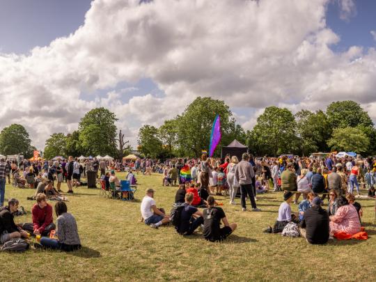 Wide shot of families sat on the grass and standing at Croydon’s Pride festival in Wandle Park in July 2023 with blue skies in the background