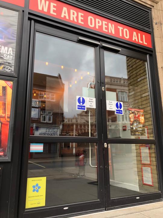 A large glass door to a public building. Sign above reads ‘We are open to all’. A big yellow sticker in the window has the Dementia Friendly Venues Charter logo