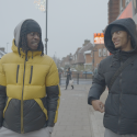 Two young men walk side by side in the street, wearing puffer ja.ckets and hoods, they look at each other and smile