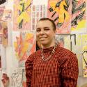A young person with short cropped hair, black and pink eye make-up and a checkered puffy shirt looks to camera, smiling. In the background, a wall covered in abstract drawings.
