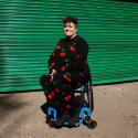 This is a picture of Jess Thom – a white woman with curly brown hair sitting in her wheelchair in front of some metallic green shop shutters on a sunny afternoon. Jess is smiling at the camera and has her hands together in her lap. She is wearing a black tracksuit with a colourful red cherry repeat print and black trainers.
